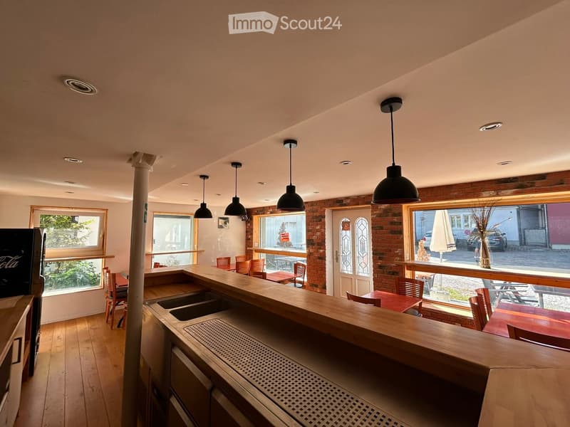 A modern bar area with a wooden counter, sink, chairs, tables, and a television, featuring large windows and exposed brick walls.