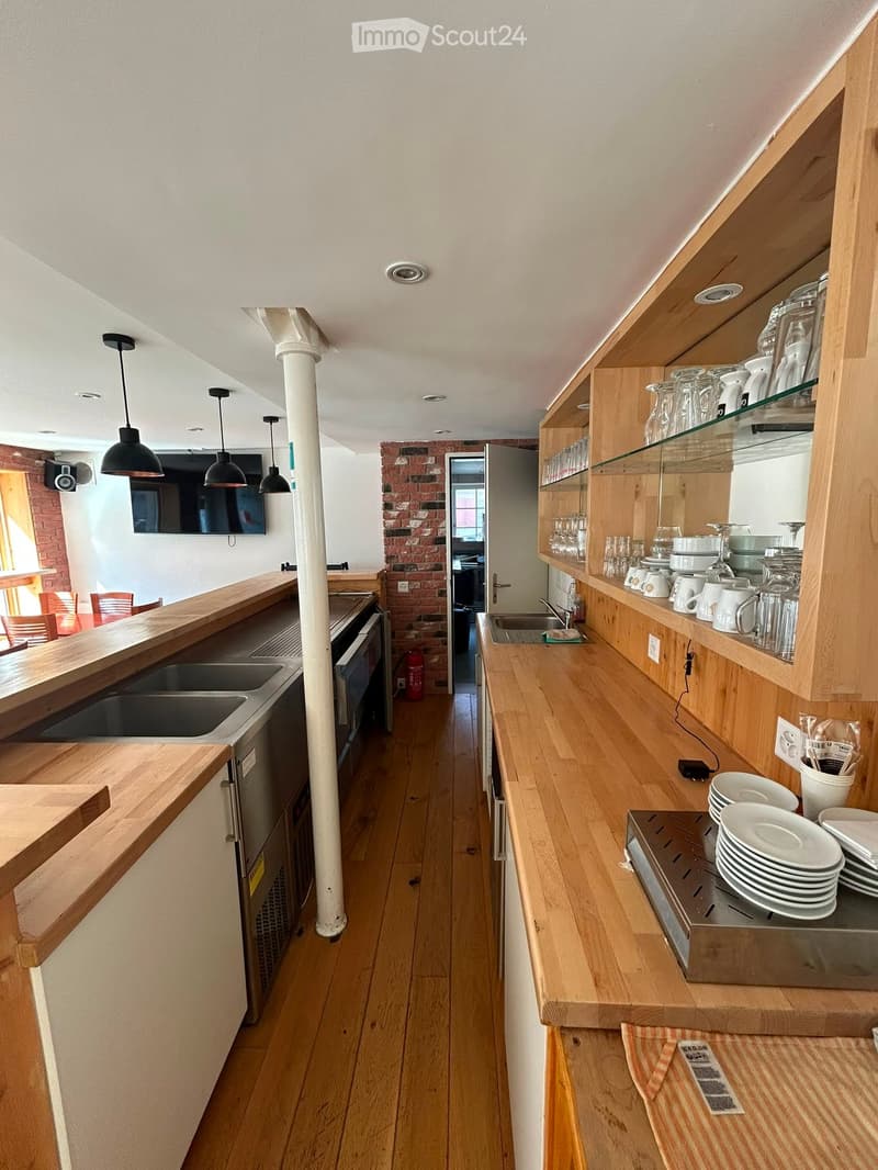Modern kitchen with wooden countertops, open shelving, sink, oven, and pendant lights. Exposed brick wall visible in the background.