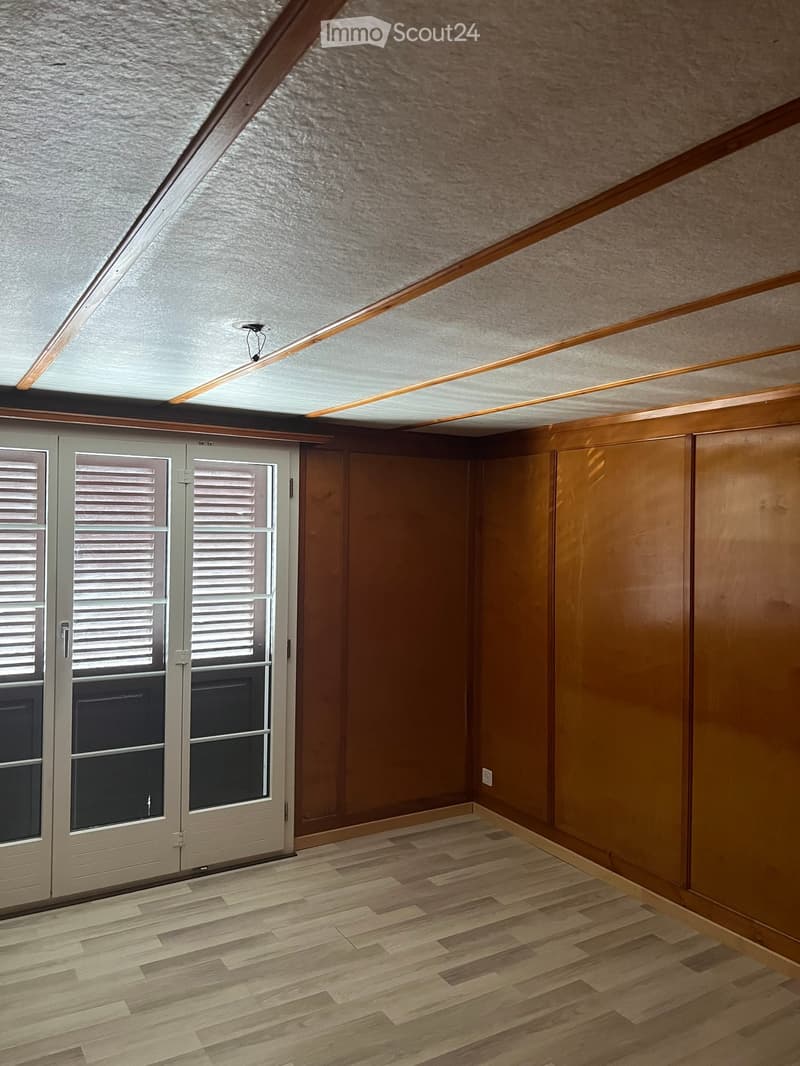 Empty room with wooden flooring, wooden walls, and a large window with shutters. The ceiling has exposed wooden beams and a skylight.