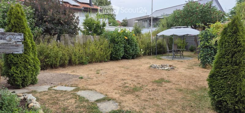 Well-maintained garden with a stone pathway, neatly trimmed hedges, a small seating area with a table and chairs under an umbrella, and a small fire pit surrounded by rocks.