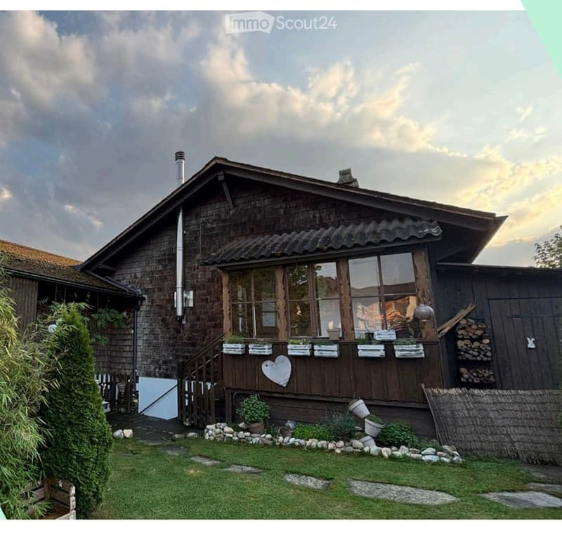 A rustic wooden house with a steeply pitched roof, large windows, and a heart-shaped wooden decoration on the facade. The house is surrounded by a small garden with potted plants and a neatly trimmed lawn.
