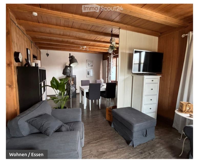 Cozy living and dining area with exposed wooden beams, a grey sofa, ottoman, dining table with chairs, television, and a fireplace.