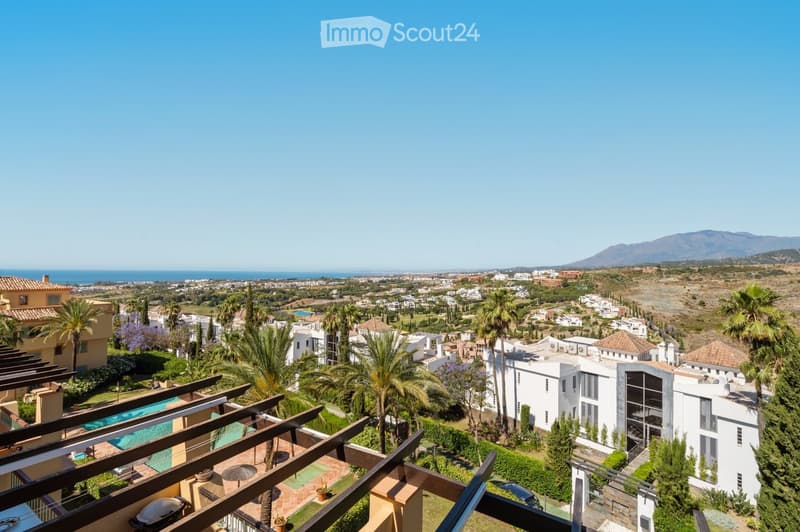A panoramic view from a balcony or terrace overlooking a coastal town with a swimming pool, palm trees, and distant mountains.