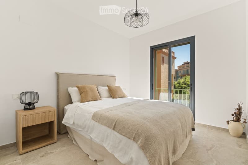 Modern bedroom with a large bed, nightstand, and decorative lamp. The room features white walls, tiled flooring, and a large window with a view of the outside.