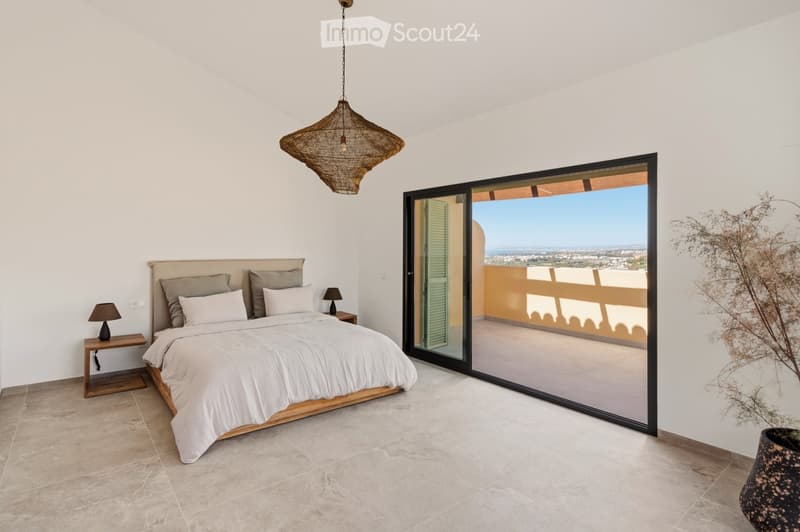Modern bedroom with a large bed, nightstands, table lamps, and a wicker pendant light. Large sliding glass doors open to a balcony with a scenic view.