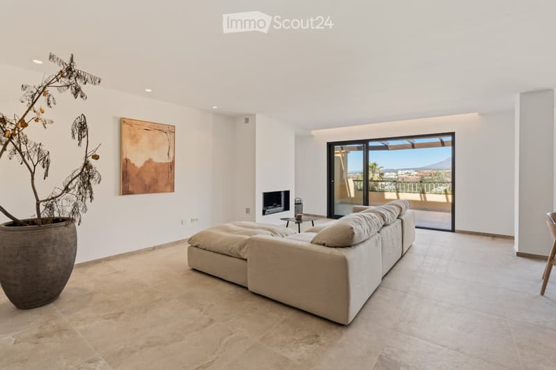 Modern living room with a large sectional sofa, fireplace, a tall decorative plant, and a view of the city from a balcony.