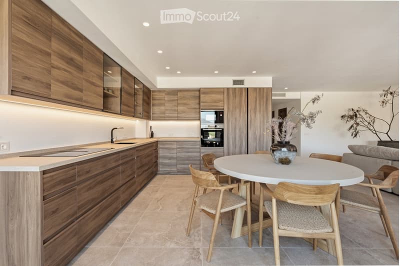 Modern kitchen with wooden cabinets, a central island, and a round dining table with chairs. The space features built-in appliances, sleek countertops, and a contemporary design.