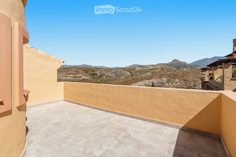 A spacious balcony with beige tiled flooring and low beige walls, offering a scenic mountain view under a clear blue sky.