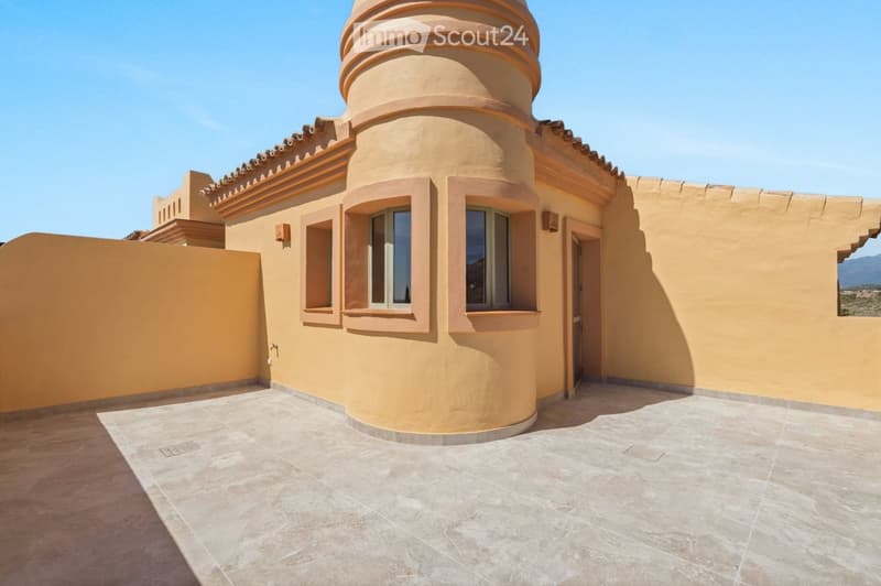 A modern, curved building with beige walls and orange accents, featuring large windows and a tiled roof, situated on a tiled patio with a clear blue sky in the background.