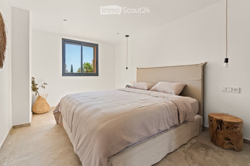 Bright bedroom with a large bed, beige bedding, a wooden stool, and a woven vase near the window offering a view of palm trees.