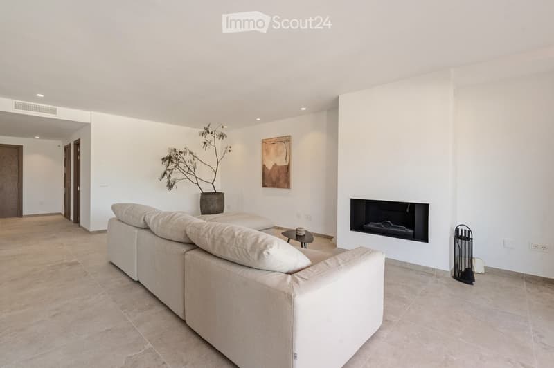 Modern living room with a beige sofa, coffee table, fireplace, and a potted plant. The room features white walls, tiled flooring, and recessed lighting.