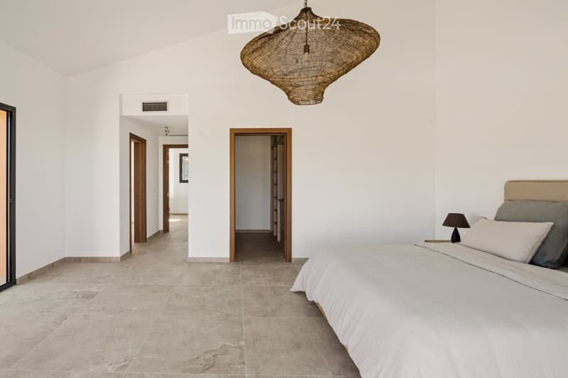 Modern bedroom with a large bed, nightstand, and a hanging woven pendant light. The room features light-colored walls, tiled flooring, and several doorways leading to other areas.