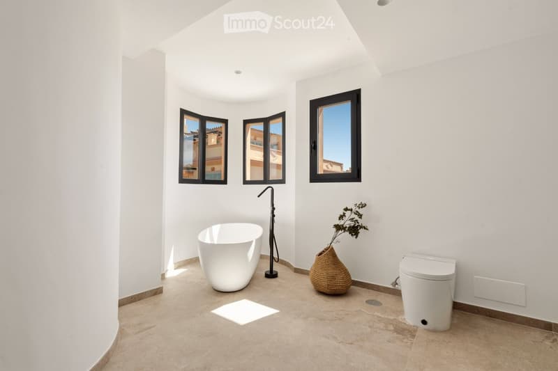 Modern bathroom with white walls, tiled floor, bathtub, toilet, and decorative vase with dried plant. Three windows provide natural light.