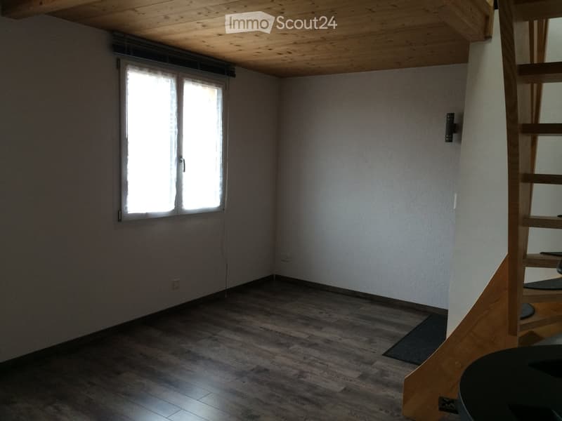 Empty room with wooden flooring, white walls, and a window letting in natural light. A wooden staircase is visible on the right side.