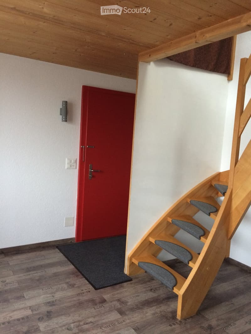 A narrow hallway with a wooden staircase leading upwards, a red door on the left, and a dark doormat in front of it. The floor is made of wooden planks with visible markings and text.