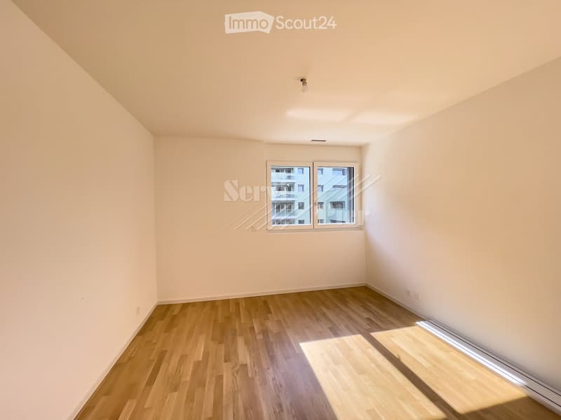 Empty room with wooden flooring, white walls, and a large window offering a view of neighboring buildings.