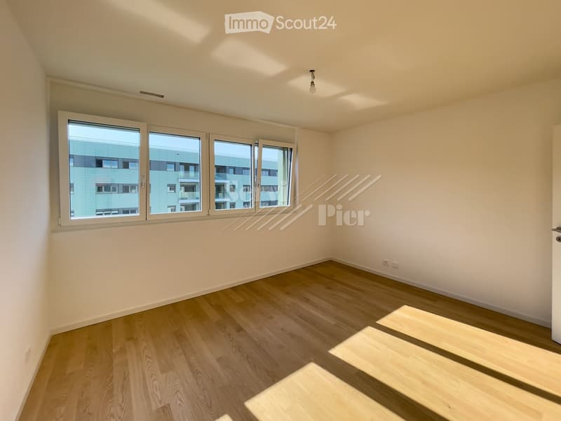 Empty room with light wooden flooring, white walls, and large windows offering a view of neighboring buildings.
