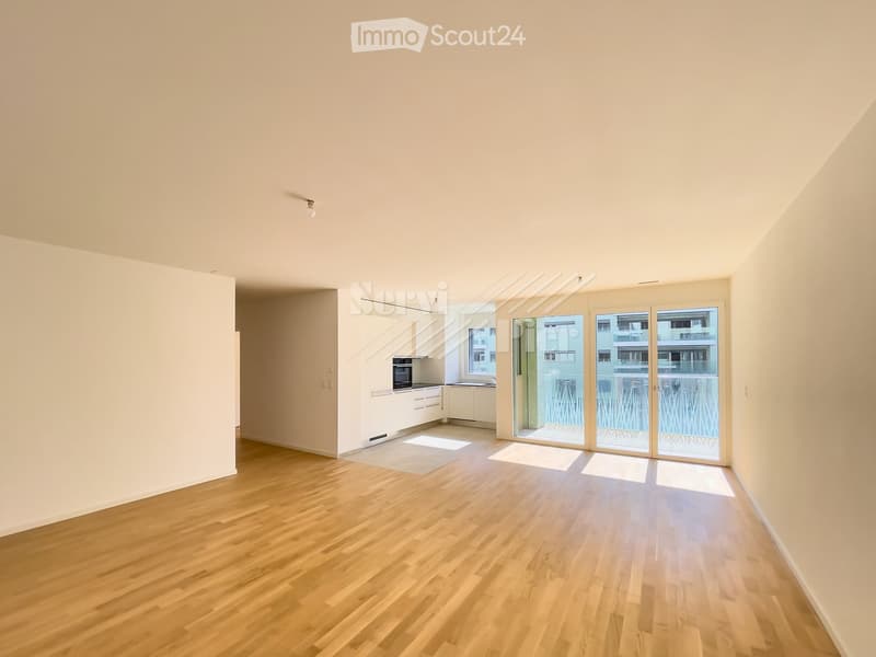 Empty room with wooden flooring, white walls, and large sliding glass doors leading to a balcony. The room is connected to a small kitchen area with white cabinets and a built-in oven.