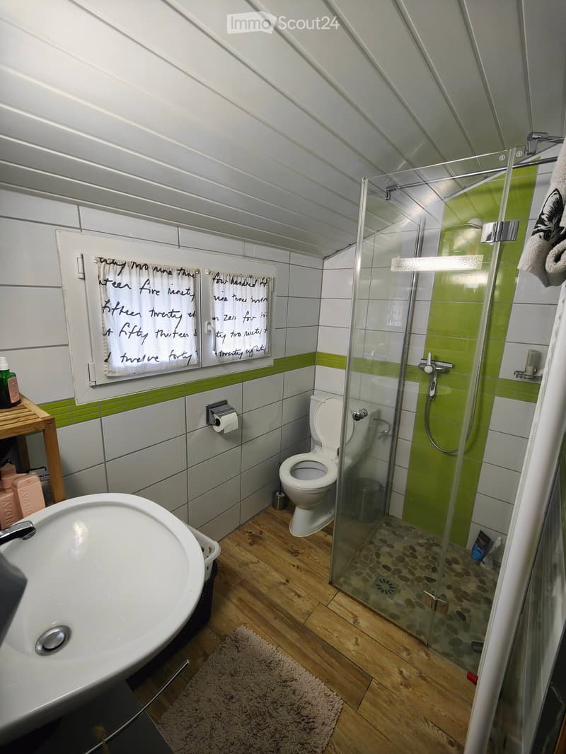 Modern bathroom with white tiles, green accents, a glass-enclosed shower, toilet, sink, and wooden shelf holding toiletries.