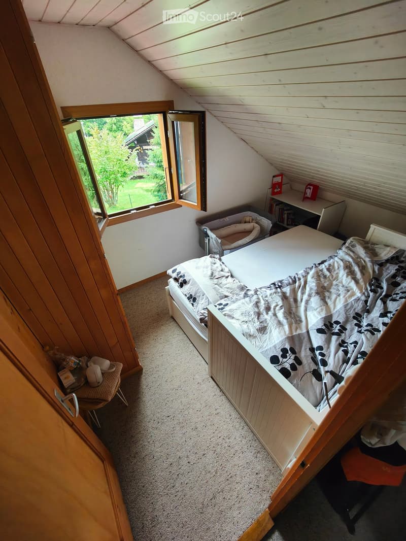 Cozy attic bedroom with a single bed, a small chair, a bookshelf, and a baby cot. Large window offers a view of the garden.