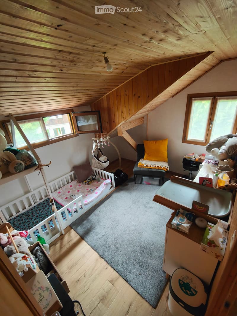 A cozy, well-lit attic room with wooden ceilings and walls, featuring a baby bed, a small table, a chair, a mirror, and various baby items.