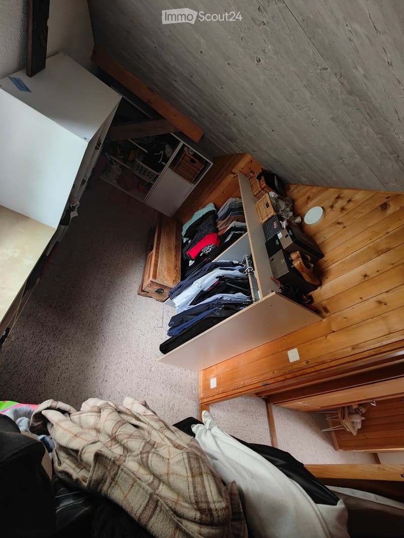 A small bedroom with a wooden wardrobe, a bed covered with a plaid blanket, and various clothing items scattered around.