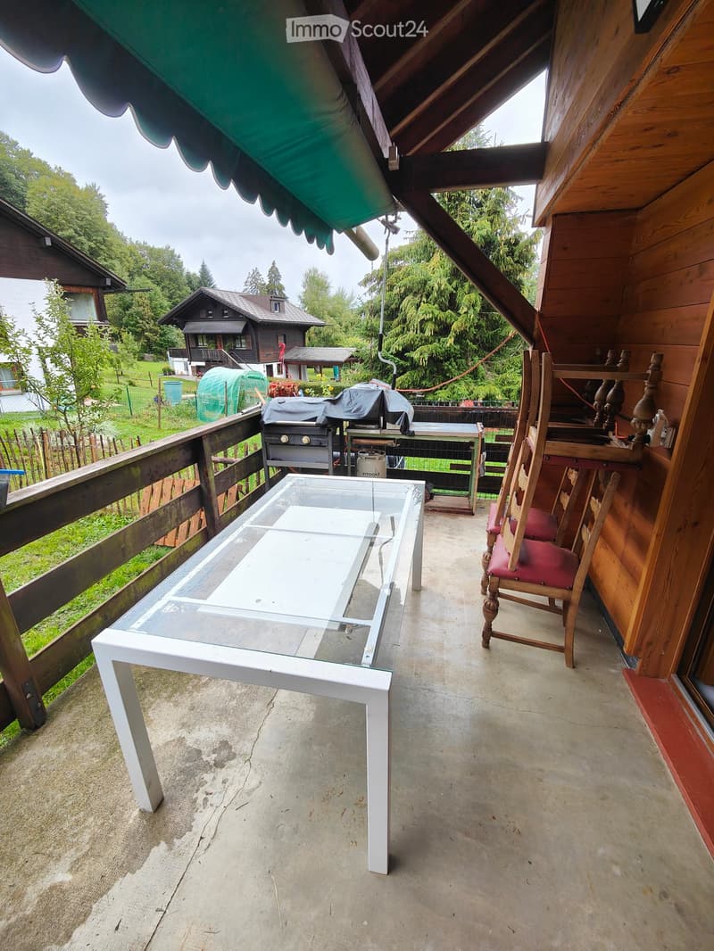 Balcony with a glass-top table and wooden chairs, overlooking a green landscape with houses and trees.