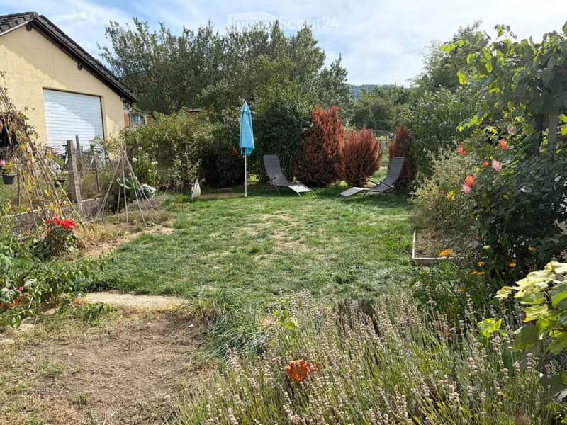 Well-maintained garden with a lawn, various plants, shrubs, and two lounge chairs near a small umbrella. A house with a garage is visible in the background.