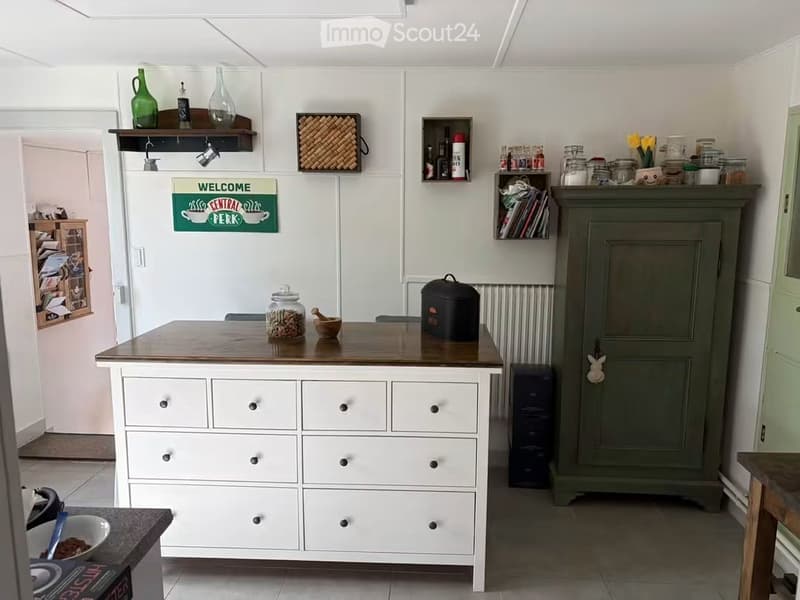 A modern kitchen with a central island, white cabinets, and various decorative items including vases, jars, and a sign that reads "Welcome Central Perk".