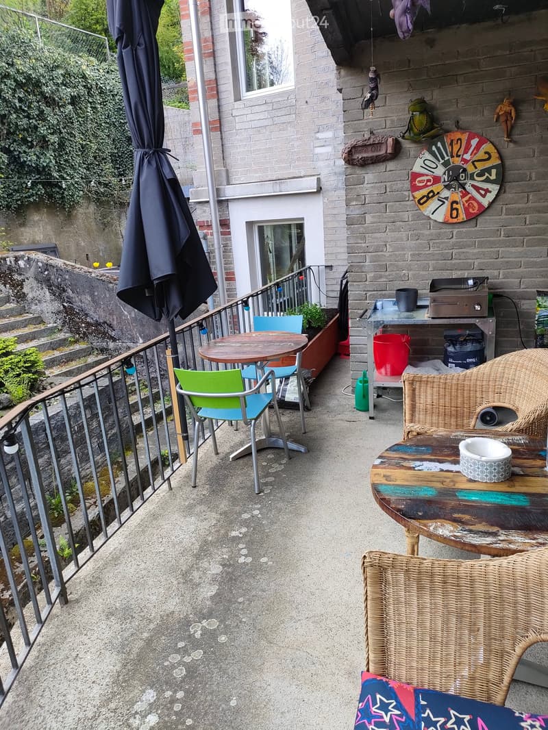 Balcony with a small seating area, including a round table, colorful chairs, and a wicker chair with a cushion. There is also a small table with a cup and a speaker, and a closed umbrella leaning against the railing.