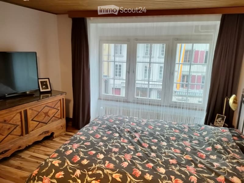 Cozy bedroom with wooden flooring, a large bed with a floral duvet, a television on a wooden stand, and a window with sheer curtains leading to a balcony.