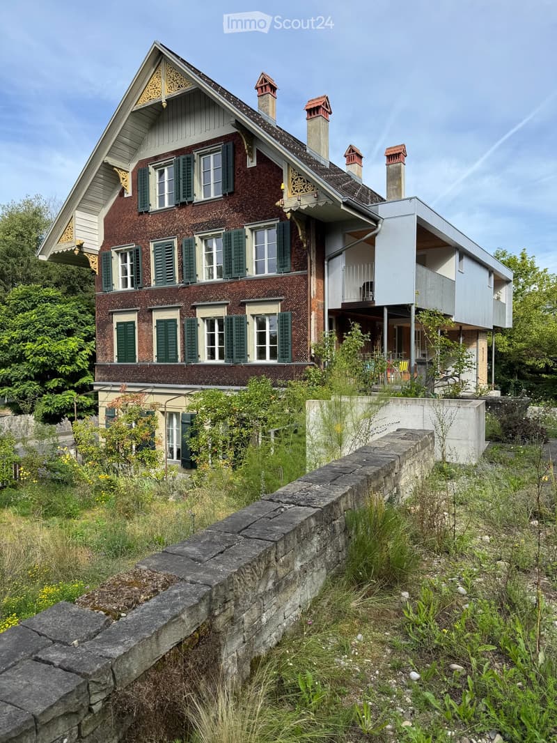 Two-story house with red brick facade, green shutters, multiple chimneys, and a modern extension with a flat roof and balcony.