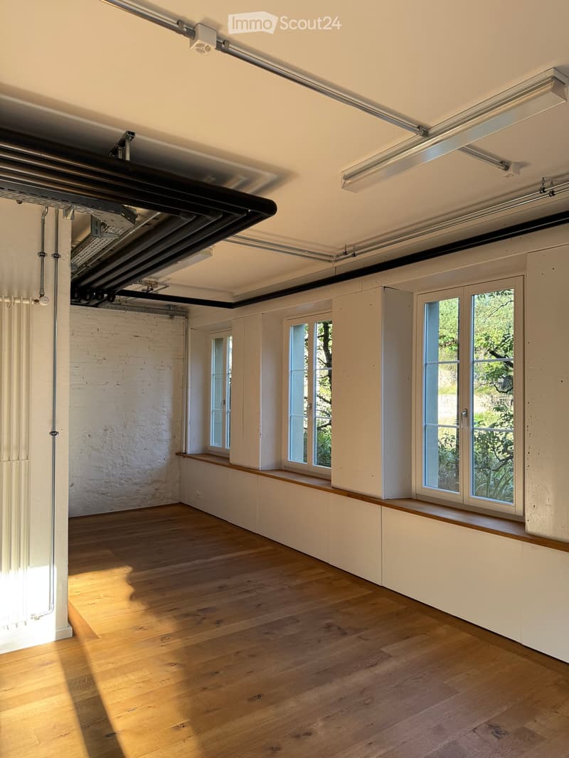 Empty room with wooden flooring, white brick wall, large windows, and built-in white cabinetry along one wall.