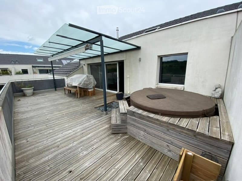 Wooden deck with a covered seating area, a hot tub, and a small bench. The deck is bordered by a low wooden railing and features potted plants.