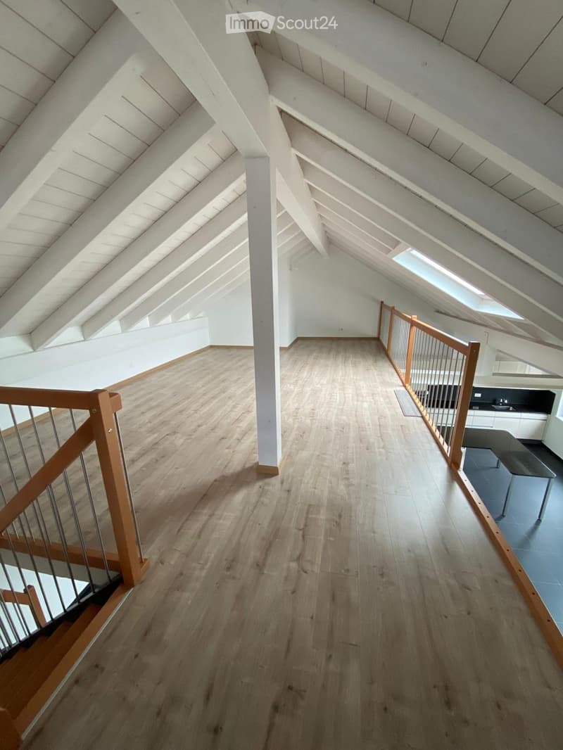 Bright, open hallway with wooden flooring, white walls, and exposed wooden beams on the ceiling. Features a wooden railing and a small kitchen area visible at the end.