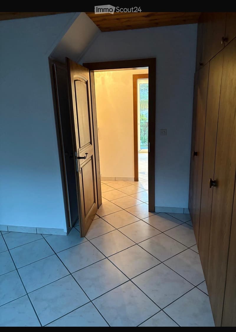 A well-lit hallway with tiled flooring, light blue walls, and wooden doors on both sides. An open doorway leads to a brightly lit room with a view of an outdoor area.