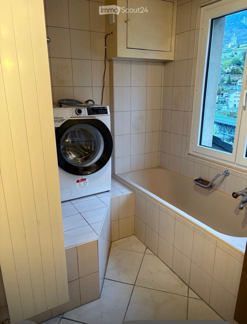 Bathroom with a bathtub, tiled walls and floor, and a washing machine in the corner. A window offers a scenic view of the town.