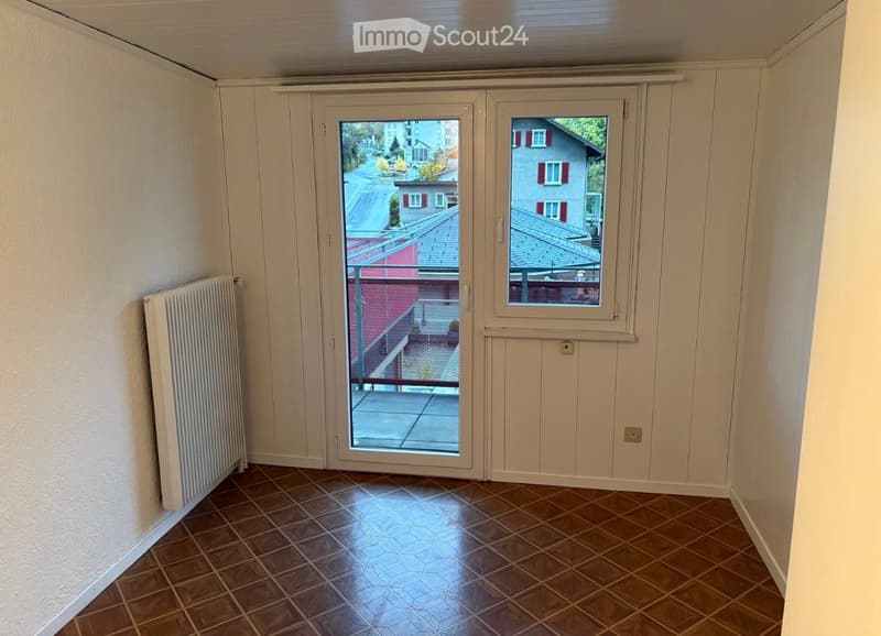 Empty room with tiled floor, white walls, radiator, and large window with a view of the street and neighboring buildings.