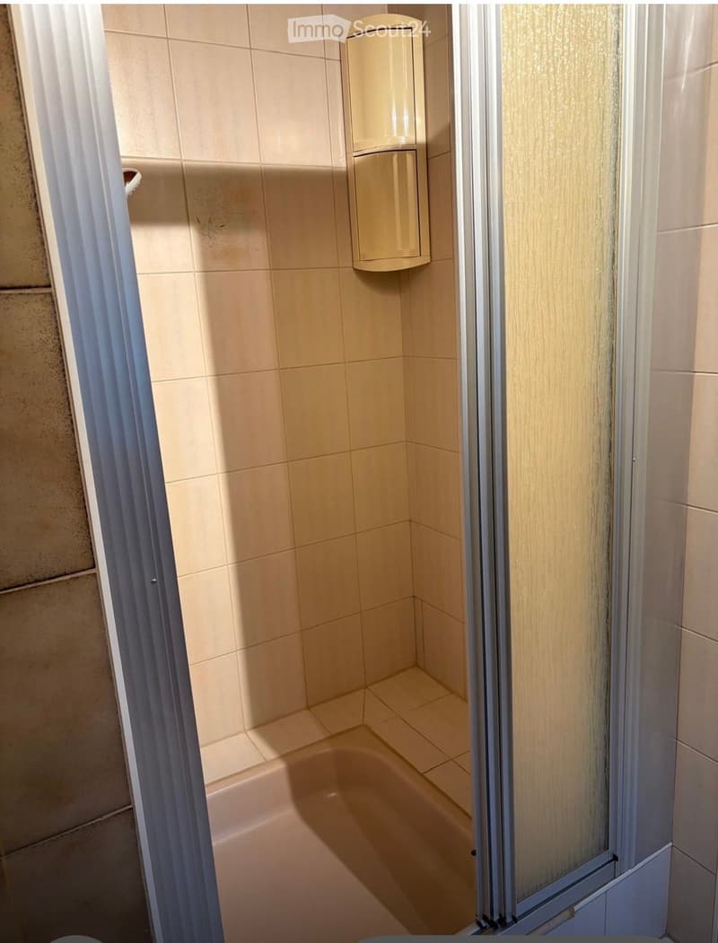 Small bathroom with beige tiled walls and floor, a toilet, a shower stall with a glass door, and a dispenser mounted on the wall.