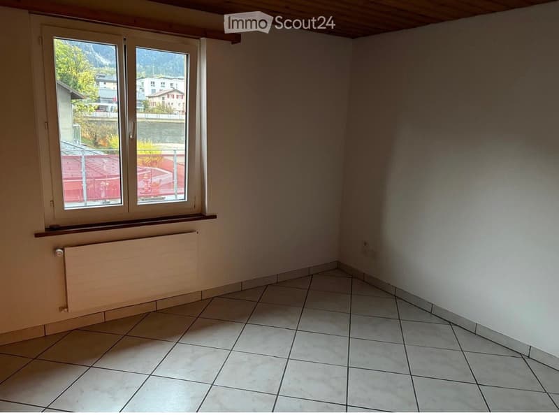 Empty room with tiled floor, white walls, large windows, radiator, and wooden ceiling beams.