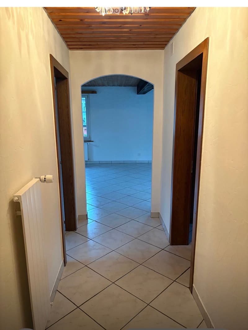 A narrow hallway with tiled flooring, wooden doors on both sides, and a wooden ceiling. An arched opening leads to another room with blue walls and a window.