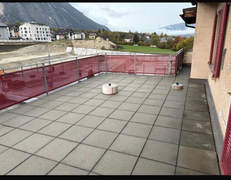 Balcony with tiled floor, metal railing, and red safety net, overlooking a construction site and mountains.