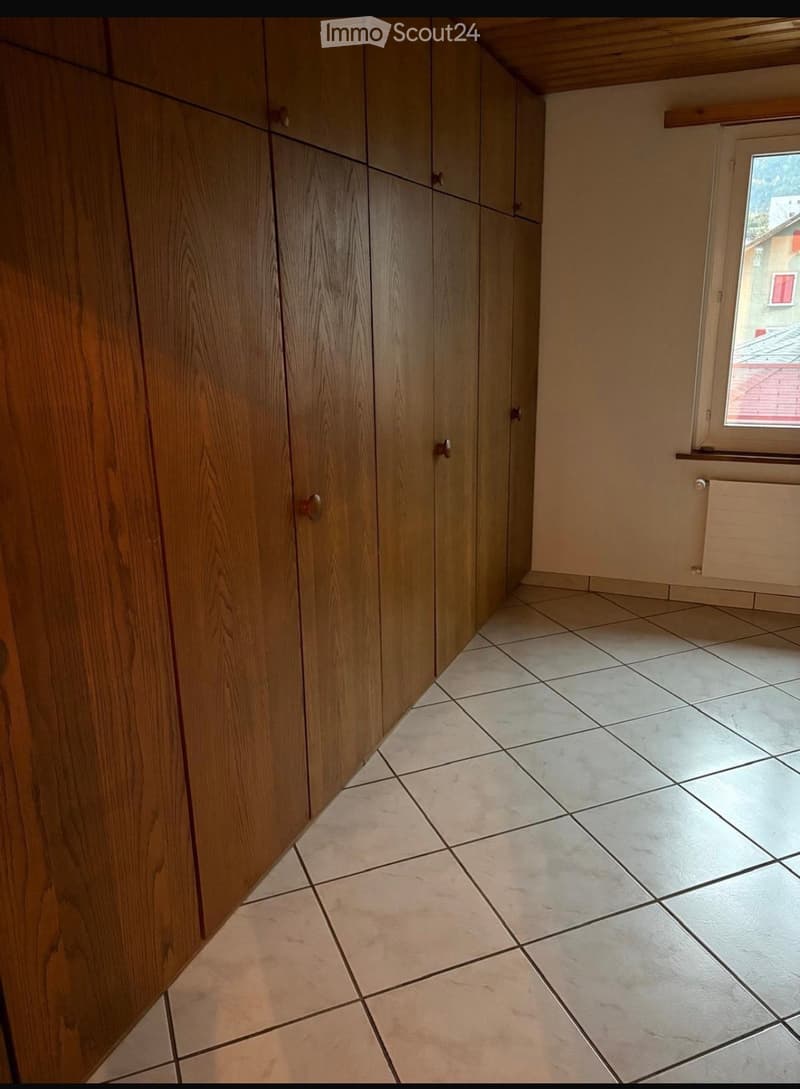 Empty room with tiled floor and wooden built-in closet, featuring a window with a view of a neighboring building.