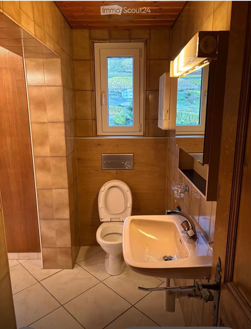 Modern bathroom with beige tiles, a toilet, a sink, and a small window offering a view of greenery.