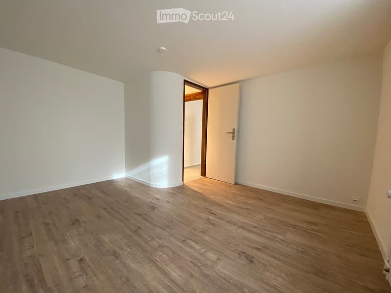 Empty room with light wooden flooring, white walls, and a central column. Two doors are visible, one leading to another room.