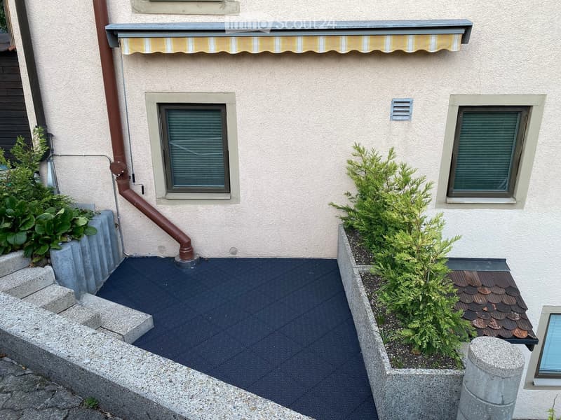 Balcony with dark flooring, small garden beds with shrubs, and a striped awning above.