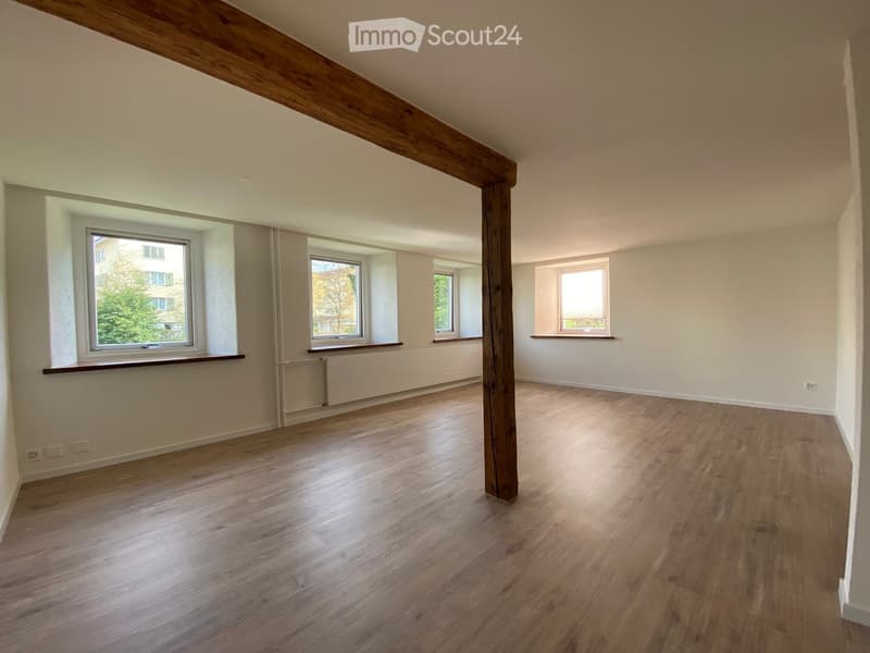 Empty room with wooden flooring, white walls, large windows, and a wooden beam in the center.