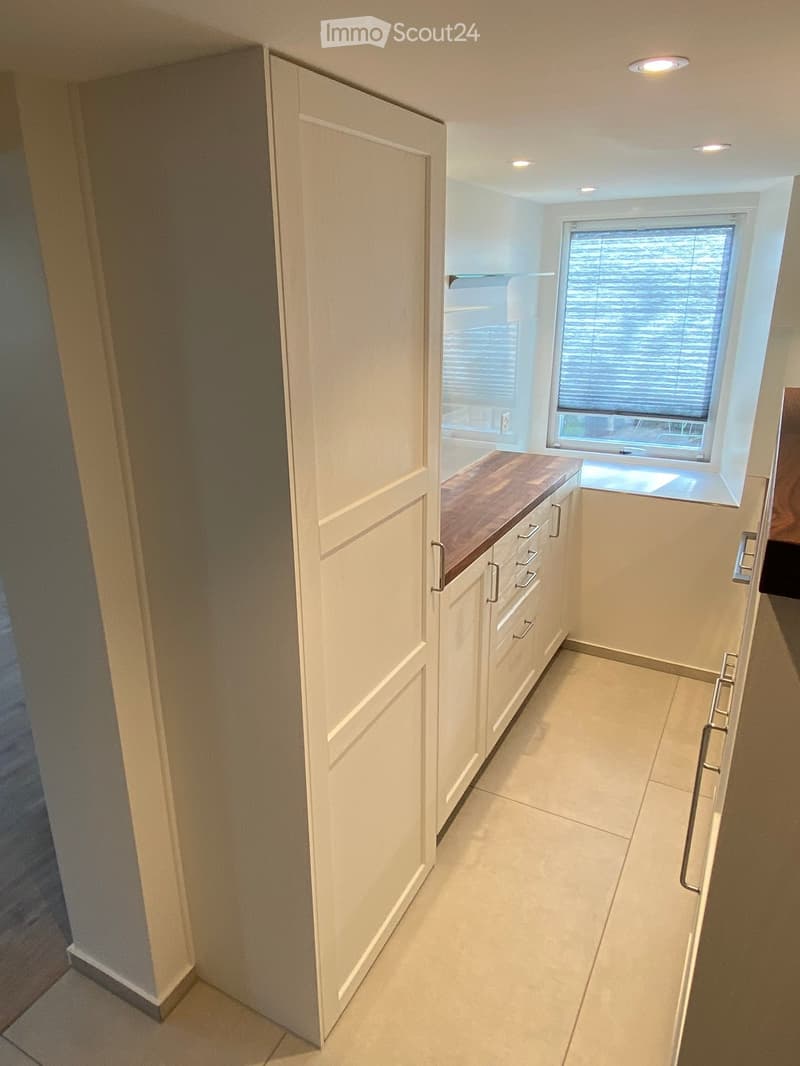 A narrow hallway with white walls, tiled floor, and built-in white cabinets with a wooden countertop and drawers. A window with blinds is visible at the end of the hallway.