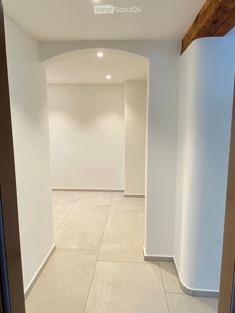 A clean, modern hallway with white walls, tiled floor, and recessed lighting. The space is well-lit and features a wooden door frame on the right side.
