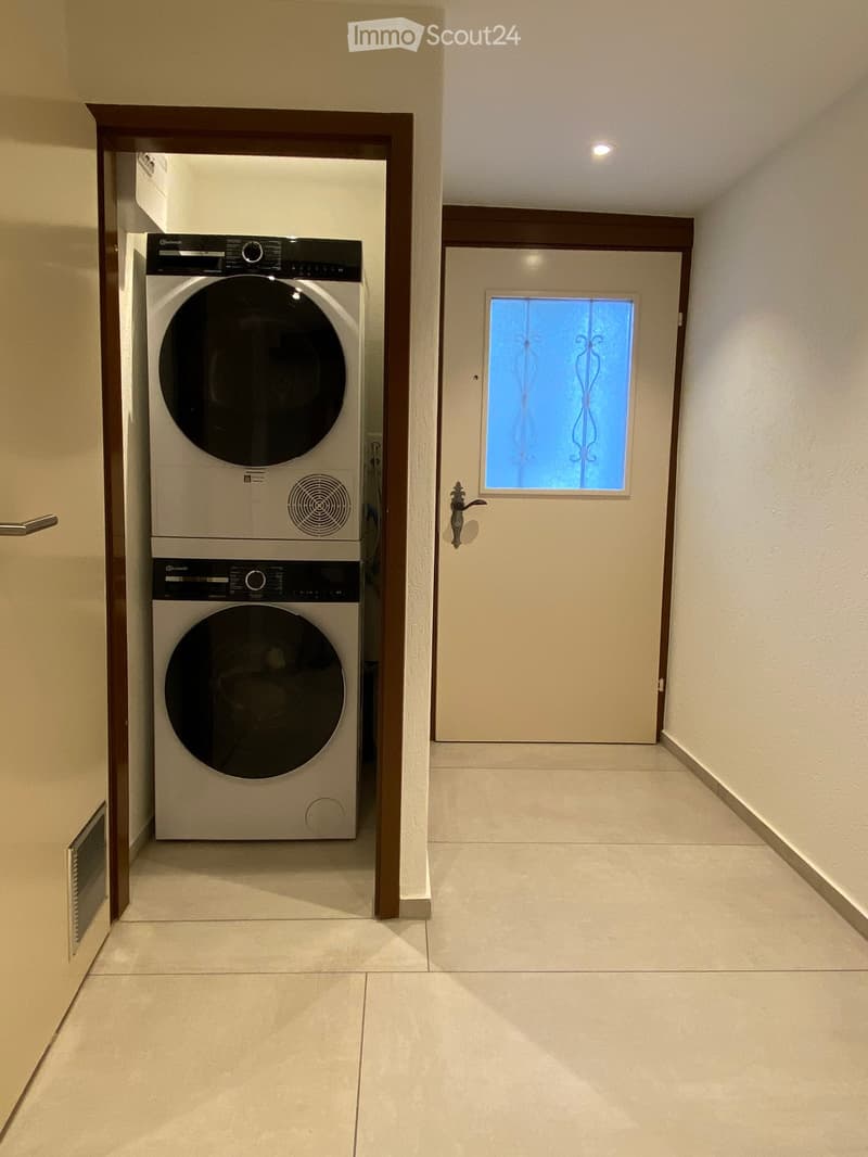 A narrow hallway with tiled flooring, a white door with a decorative window, and a stacked washer and dryer unit on the left side.